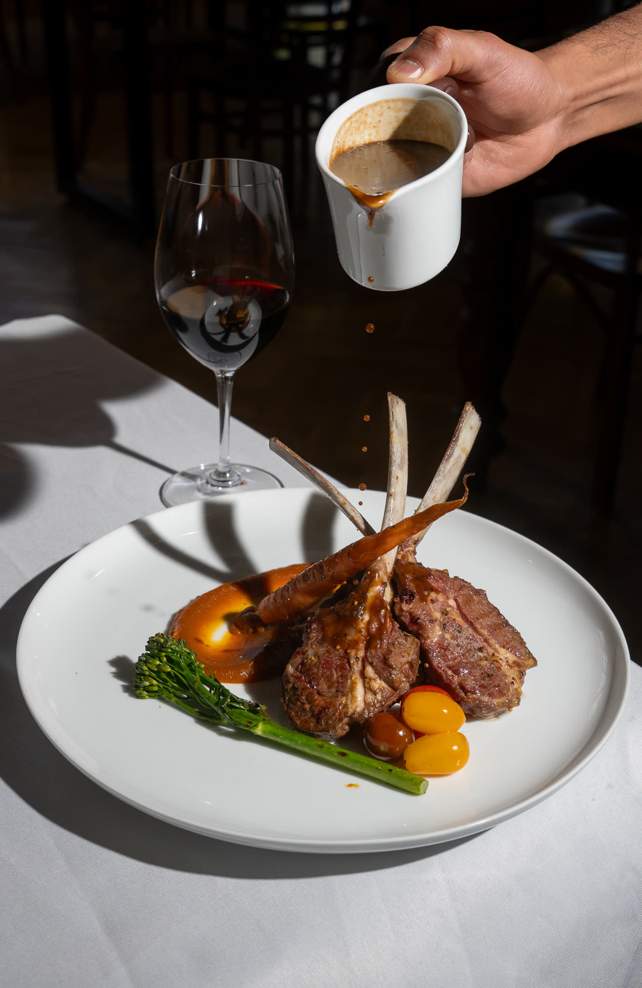 Gravy being poured over plated lamb cutlets with carrot and broccolini, beside a glass of red wine.