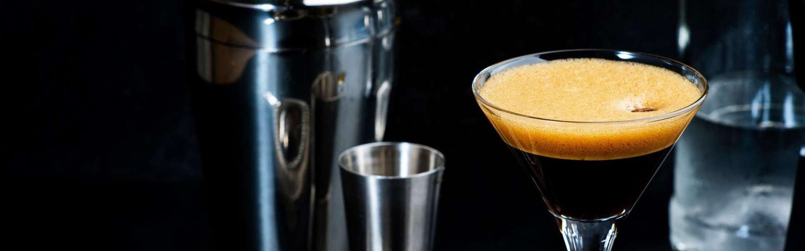 An espresso martini with a cocktail shaker and jigger behind it.